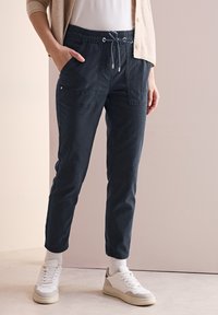 Marinblå tapered byxor med elastisk midja, dragsko, två framfickor och manschetter, kombinerat med vita sneakers.