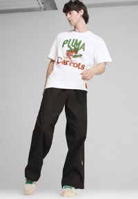 Balta T-kreka ar zaļu un oranžu "PUMA Carrots" grafiku, apvienota ar melnām platām biksēm un bēšām kedām ar zaļiem akcentiem.
