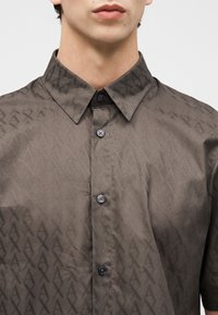 Κοντινό πλάνο ενός ατόμου που φορά γκρι πουκάμισο με κουμπιά, με μυτερό γιακά και επαναλαμβανόμενο γεωμετρικό σχέδιο.