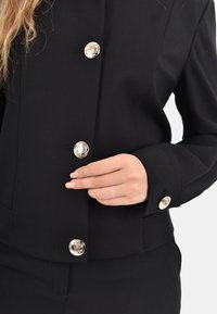 Blazer nero aderente realizzato in tessuto elasticizzato, con bottoni d'argento, design sartoriale e texture liscia. Primo piano dei bottoni e della manica.