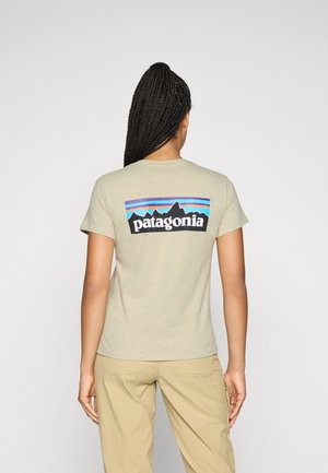 Vrouw met lange gevlochten haren draagt een beige Patagonia T-shirt en khaki broek, zijwaarts gekeerd tegen een effen lichte achtergrond.
