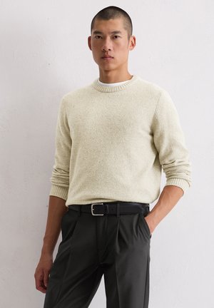 Lichtbeige trui met een gestructureerde afwerking, ronde halslijn en geribbelde manchetten, in combinatie met zwarte broek en een zwarte riem.