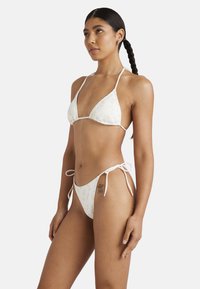 Bikini triangolo in tessuto bianco con motivo floreale; presenta spalline sottili regolabili e slip bikini con lacci laterali. Texture morbida.