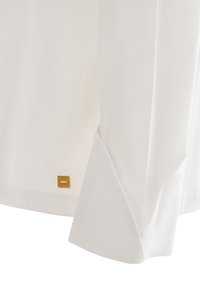 Camicetta bianca a maniche lunghe con una texture liscia e un'etichetta del marchio dorata sull'orlo. I polsini sono leggermente arrotolati con un design a taglio dritto.