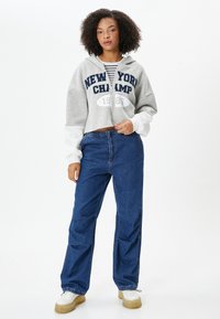 Graue, kurze Kapuzenjacke mit "NEW YORK CHAMP 1985" in Marineblau bedruckt. Kombiniert mit blauen, lässigen Jeans und weißen Sneakers mit beigen Sohlen.