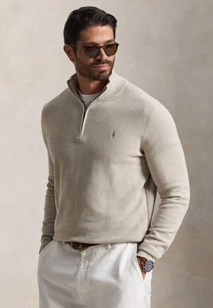 Mand iført beige kvart-zip sweater, hvide bukser, solbriller og armbåndsur, stående med hænderne i lommerne mod en ensfarvet baggrund.