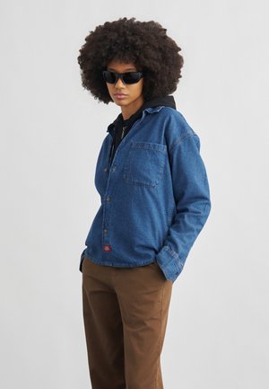 Person med krøllet hår iført sorte solbriller, blå denimjakke over en sort hættetrøje og brune bukser, står med hænderne i lommerne.