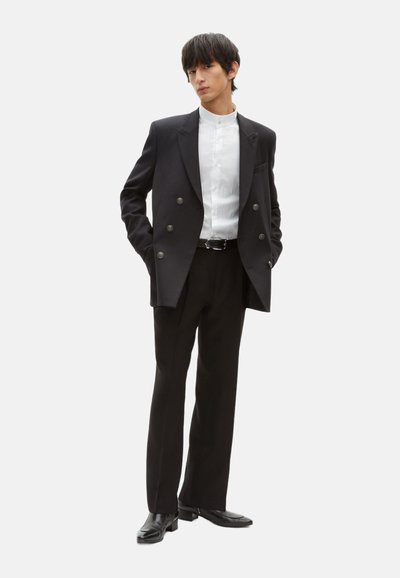 Schwarzes, doppelt geknöpftes Blazer mit silbernen Knöpfen, weißes Hemd mit Kragen, schwarze Hose und schwarze Schuhe, vor einem weißen Hintergrund stehend.