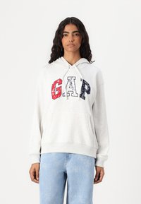 Hoodie gris clair avec une poche kangourou, portant "GAP" en lettres rouges, blanches et bleues à motif ; poignets et ourlet côtelés.