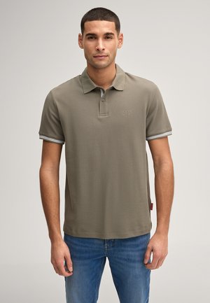 Uomo in piedi che indossa una polo verde oliva a maniche corte con polsini a righe bianche e jeans blu su sfondo neutro.