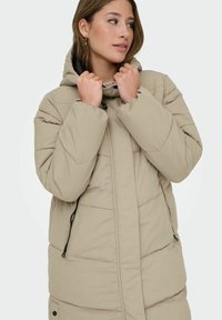 Chaqueta acolchada beige con capucha, que presenta bolsillos con cremallera, diseño acolchado y una textura exterior suave. Estilo minimalista con herrajes discretos.