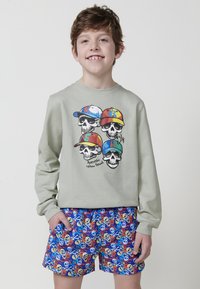 Sudadera gris con gráfico de calavera que lleva gorras coloridas, combinada con pantalones cortos azules con un patrón de calavera pirata y huesos cruzados.