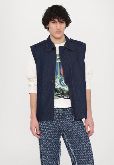 Denim vest with a collar, dark blue color, front button closure. Layered over a graphic print sweatshirt and distressed patterned denim pants.