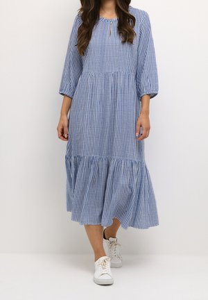 Kvinde iført midi-kjole med blå og hvide lodrette striber, 3/4-ærmer og hvide sneakers, stående foran en ensfarvet baggrund.