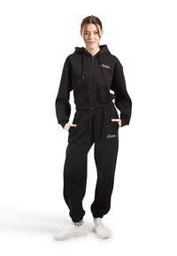 Zwarte cropped hoodie met rits en voorzakken, gecombineerd met zwarte joggerbroeken met een geborduurd logo. Witte sneakers maken de outfit compleet.