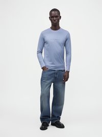 Άνδρας που φοράει ανοιχτό μπλε μακρυμάνικο πουκάμισο, φαρδιά μπλε τζιν και μαύρα παπούτσια, στέκεται με το ένα χέρι στην τσέπη σε απλό λευκό φόντο.