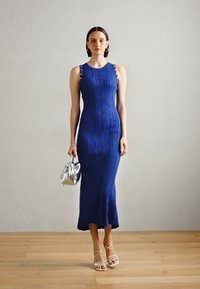 Robe midi bleue à côtes avec une quincaillerie d'épaule, associée à un sac à main argenté. Portée avec des sandales à talons blancs à lanières sur un sol en bois.