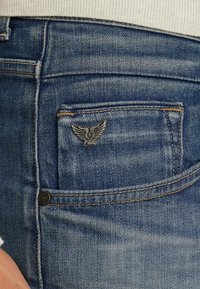 Jeans de mezclilla azul con una textura descolorida, que presentan un pequeño emblema plateado con alas en el bolsillo trasero y costuras naranjas contrastantes.