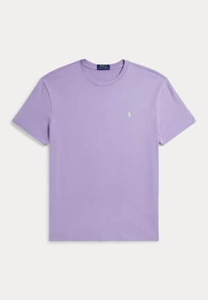 Homme portant une chemise à manches courtes violet clair, un pantalon blanc plissé, des mocassins marron et une montre-bracelet, debout devant un fond neutre.