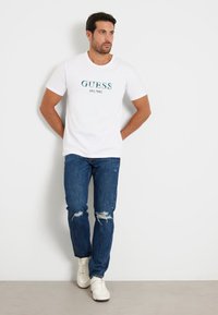 T-shirt en coton blanc avec un graphique bleu "GUESS EST. 1981", associé à un jean bleu foncé avec des détails déchirés et des baskets blanches.