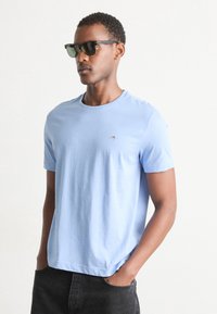 Light blue cotton t-shirt with short sleeves and a crew neckline. Features a small logo on the chest. Worn with dark jeans and sunglasses.