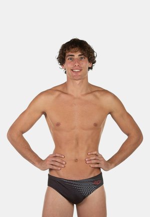 Giovane uomo con capelli ricci in piedi con sicurezza con le mani sui fianchi, indossa slip da bagno neri con fantasia su sfondo semplice.