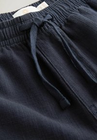 Pantalone s elastičnim pojasom u mornarsko plavoj boji izrađeni su od teksturirane tkanine i imaju vezicu te ležeran kroj. Prikaz detalja naglašava mekoću tkanine.