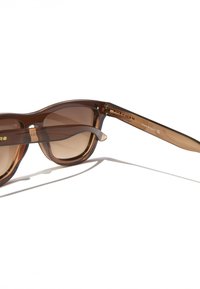 Barna műanyag napszemüveg árnyalatos lencsékkel, lekerekített formával és keskenyedő szárakkal. Márkajelzéssel a szemöldök felett és világos, áttetsző kerettel rendelkezik.