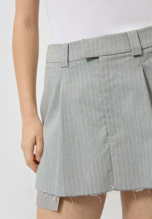 Femme portant une jupe gris clair à fines rayures avec un ourlet effiloché et un haut blanc sans manches en côtes, main reposant le long du corps.