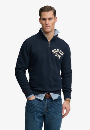 Mand iført mørkeblå kvart-lynlåstrøje med teksten "Super Dry", lagdelt over en lyseblå skjorte, kombineret med blå jeans og et brunt bælte.