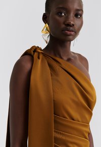 Vestido de cetim castanho com um ombro drapeado e detalhe de nó. Modelo usa brincos grandes triangulares em ouro e prata. Textura suave e design ajustado.