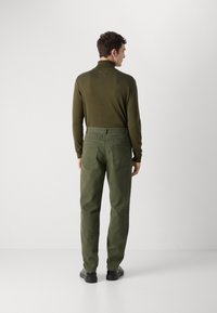 Olive green, long-sleeve shirt with high collar paired with loose-fit, high-waisted olive green trousers featuring back pockets and black shoes.