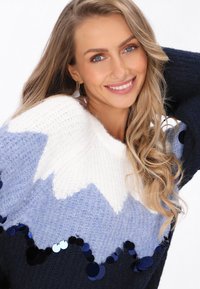 Niebieski i biały sweter dziewiarski z wzorem w zygzak, zdobiony ciemnymi cekinami wzdłuż dołu; miękka tekstura i okrągły dekolt.