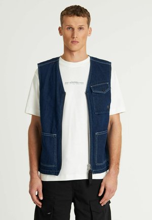 Denim ærmeløs vest, mørkeblå, lynlås foran, brystlomme med trykknap, to nederste lommer, hvid skjorte under, båret med sorte cargo bukser.