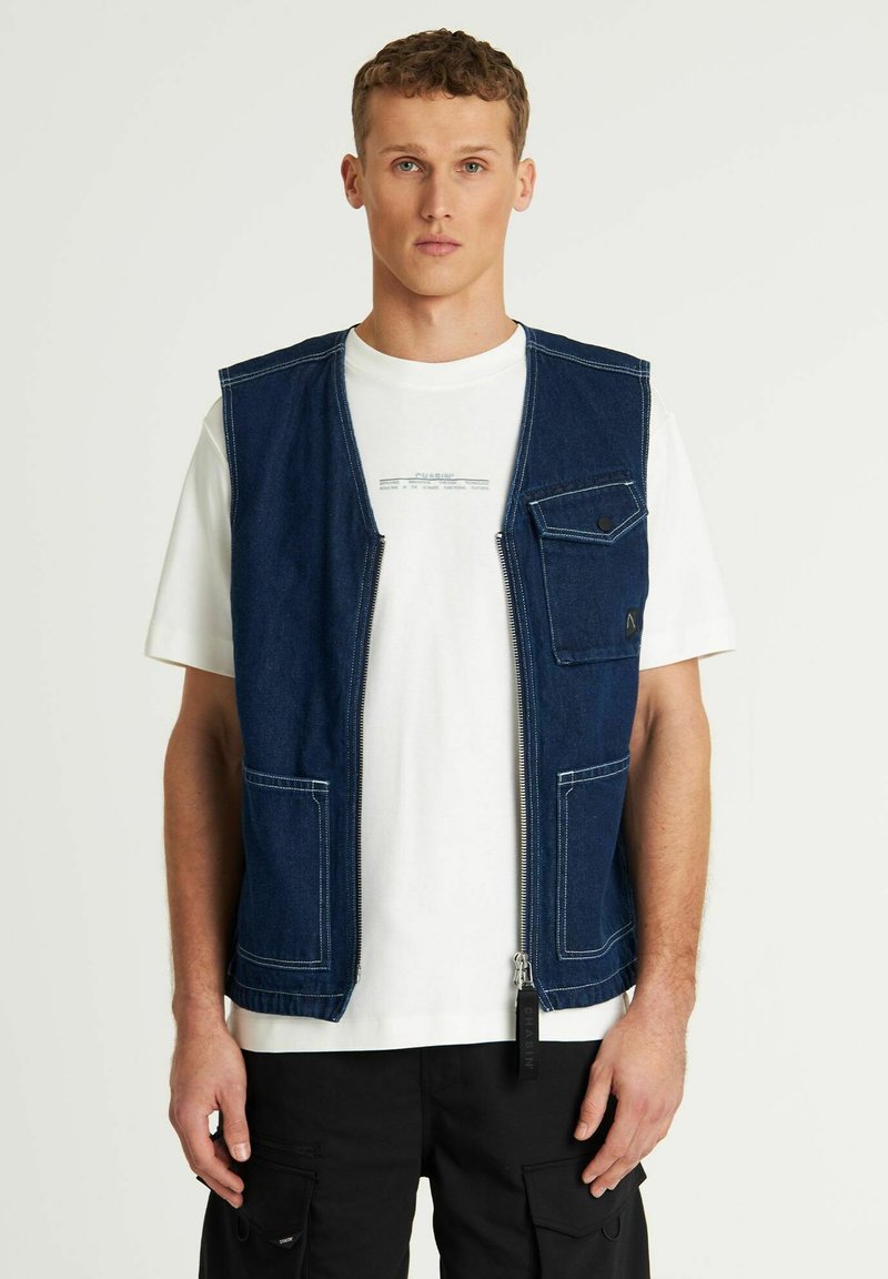 Denim brezrokavnik, temno moder, z zadrgo spredaj, žep na prsih s pritrdilnikom, dva spodnja žepa, bela srajca pod njim, nosi se z črnimi cargo hlačami.