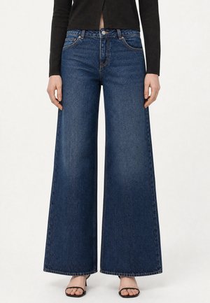 Personne portant un jean bleu foncé à jambes larges, un haut noir à manches longues et des talons ouverts noirs, debout devant un fond blanc.