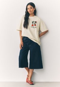 Krém színű, túlméretezett póló paradicsomos grafikával, sötét culotte farmerrel és piros Mary Jane cipővel, világos fal előtt állva.