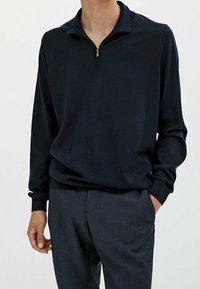 Marineblaue Zip-up-Sweatjacke aus weichem Material, mit Stehkragen und langen Ärmeln, kombiniert mit maßgeschneiderten dunklen Hosen.