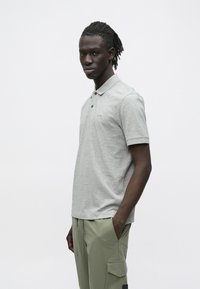 Noor mustanahaga mees, kellel on patsidega juuksed, kandes helehallidest polosärki ja lahtisi helerohelisi pükse, valge tausta ees.