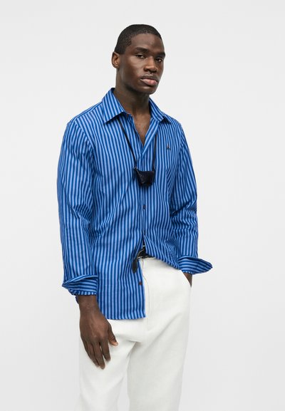 Νέος άνδρας που φοράει μπλε ριγέ μακρυμάνικο πουκάμισο, λευκό παντελόνι και μια μικρή μαύρη θήκη κρεμασμένη από κορδόνι γύρω από το λαιμό του.