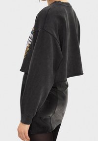 Avskuren svart sweatshirt med oversized ärmar, prydd med färgglad grafisk broderi på sidan. Bärs med en svart läder minikjol.