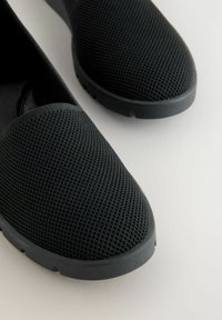 Para czarnych butów na slip-on z oddychającymi siateczkowymi cholewkami i grubymi gumowymi podeszwami na białej powierzchni.