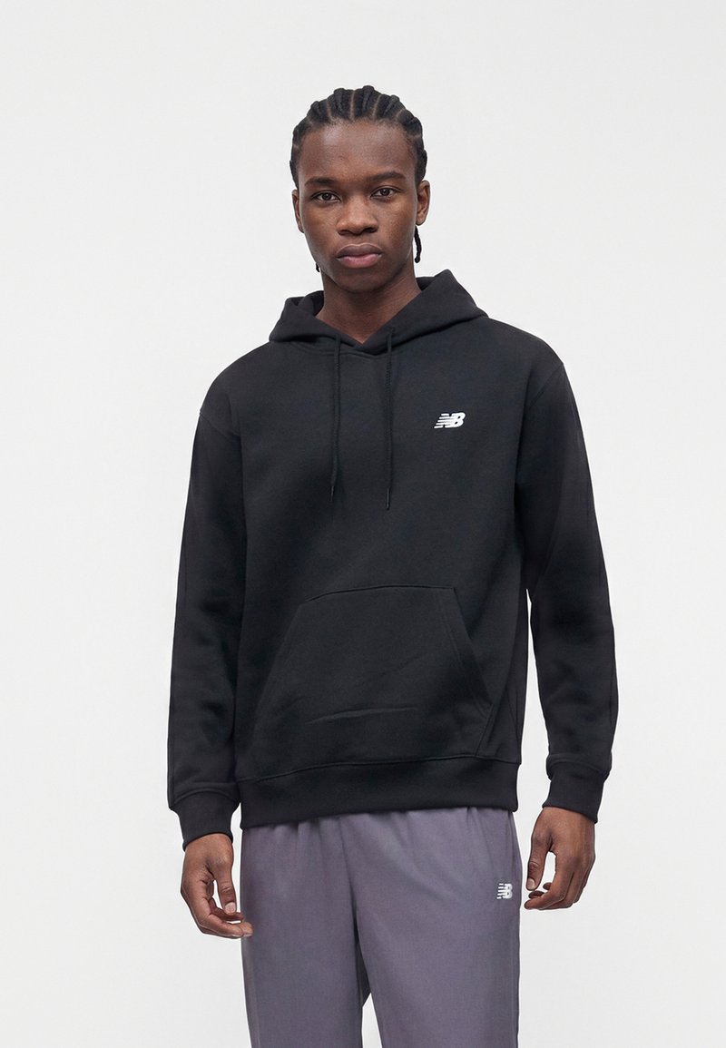 Ung mann med flettet hår som har på seg en svart hettegenser og grå joggebukser, begge med hvite New Balance-logoer, foran en ensfarget bakgrunn.