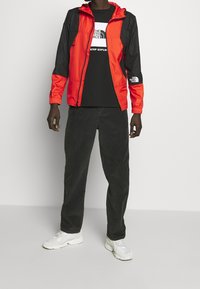 Červeno-černá větrovka s kapucí, zapínáním na zip a s logem, doplněná černým tričkem a tmavými manšestrovými kalhotami. Bílé tenisky.