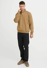 Brown zip-neck sweater with ribbed cuffs and hem, paired with black trousers and beige shoes, standing against a plain background.