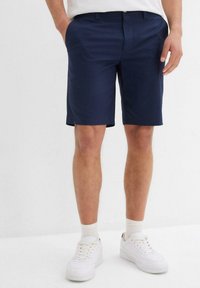 Pantaloni corti blu navy realizzati in tessuto liscio, con una vestibilità su misura, davanti piatto, passanti per la cintura e tasche laterali. Abbinati a sneakers bianche.