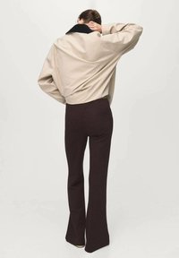 Personne portant une veste oversized beige avec un col noir et un pantalon évasé marron foncé, se tenant de dos à la caméra, la main sur le cou.