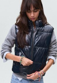 Marinblå puffy väst med hög krage och glänsande yta, kombinerad med en grå tröja. Anmärkningsvärda detaljer inkluderar flera färgglada armband och en dragkedja.