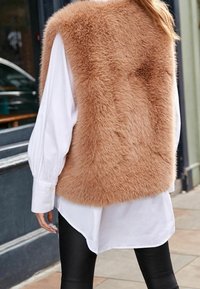 Gilet en fausse fourrure de couleur marron doux, à col haut, superposé sur une chemise blanche à boutons. Un legging noir complète la tenue. Texture lisse et volume.