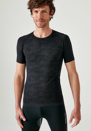 Man met warrig bruin haar, draagt een nauwsluitend zwart sportshirt met korte mouwen en zwarte leggings met witte tekst op de dij.